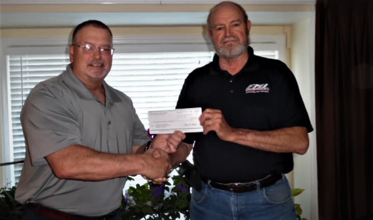 Two men shaking hands and holding a check together in front of a window.